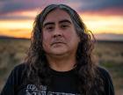 Portrait of artist Raven Chacon wearing a black tee shirt with white graphic, standing in an arid field with the sun low on the horizon.